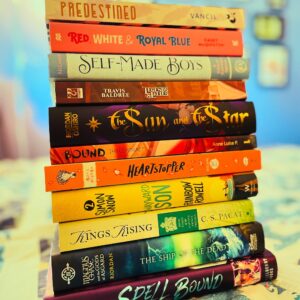 ‘An expanded Pride flag stack of 11 queer books | Predestined, Red, White, and Royal Blue, Self-Made Boys, Legends and Lattes, The Sun and The Star, Bound The Contract, Heartstopper, Kings Rising, Wayward Son, Magnus Chase and the Gods of Asgard & Spell Bound’ — “11 Books You Should Add to Your Rainbow Stack this Pride”, Kat Vancil, The Saga