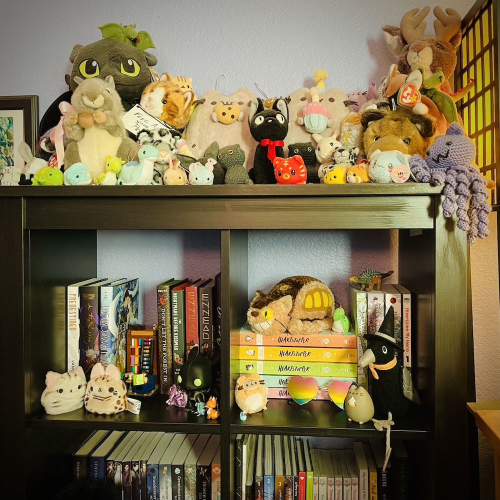 Kat Vancil’s short bookcase within her story studio featuring over 50 mini plush, a selection of Queer books, and a limited edition LEGO bookcase bookend